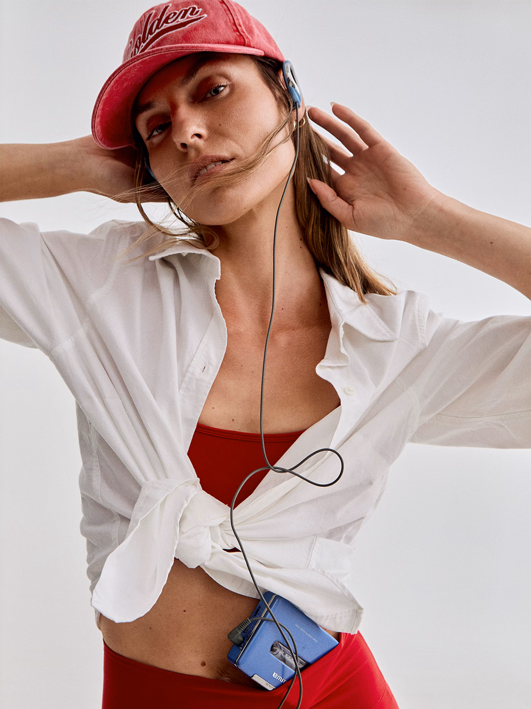 Model in a red Deva sports bra and shorts under a knotted white shirt, wearing a red cap and listening to a cassette player.